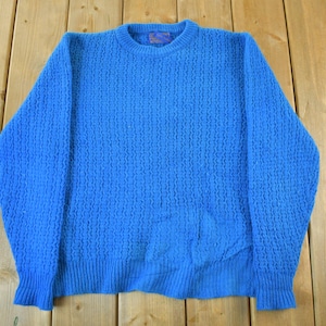 May include: A bright blue crew neck sweater with a textured pattern. The sweater has long sleeves and a ribbed hem and cuffs. The garment is made of a soft material and is displayed on a wooden surface.