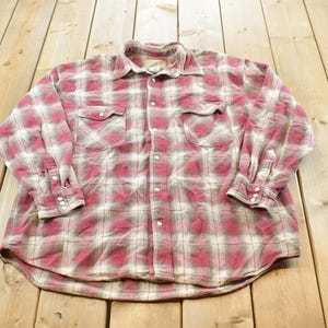 May include: A long-sleeve button-down shirt featuring a plaid pattern in shades of red, white, and grey. The shirt includes a standard collar, two chest pockets with buttoned flaps, and buttoned cuffs. The front is secured with a series of white buttons.