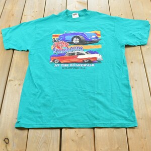 May include: A teal short-sleeve t-shirt featuring a graphic of two classic cars. The text reads Cruisin The Wildwoods AT THE BOARDWALK WILDWOOD N.J. The top car is blue and the bottom car is red and white with flames on the side panels.