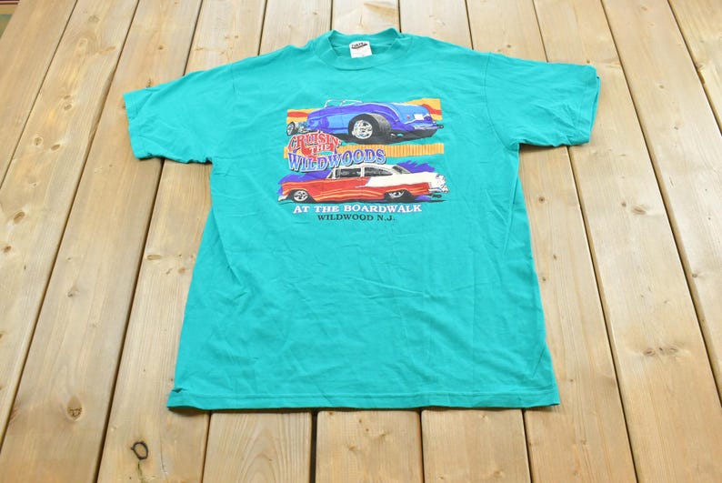 May include: A teal short-sleeve t-shirt featuring a graphic of two classic cars. The text reads Cruisin The Wildwoods AT THE BOARDWALK WILDWOOD N.J. The top car is blue and the bottom car is red and white with flames on the side panels.