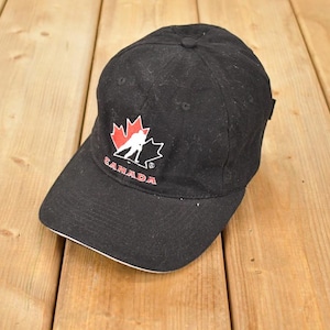 Peut inclure: Casquette de baseball noire avec une bordure blanche. Le chapeau présente un logo de feuille d'érable rouge avec une silhouette de joueur de hockey et le mot "CANADA" en rouge. La casquette est sur une surface en bois.