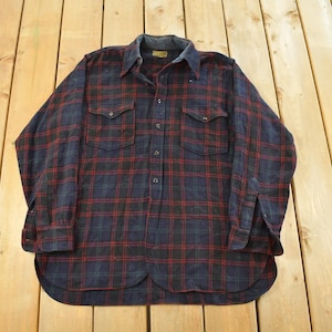 May include: A long-sleeved, button-down shirt in a blue and red plaid pattern. The shirt has two chest pockets and a collar. The fabric appears to be a flannel or similar material. The shirt is laid flat on a wooden surface.