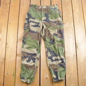 May include: Camouflage cargo trousers with a drawstring at the bottom of the legs. The trousers are a dark green, brown, and black camouflage pattern.