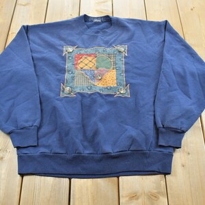 Peut inclure: Un sweat-shirt bleu marine à manches longues avec un empiècement carré central au motif patchwork multicolore. L'empiècement est encadré par une bordure décorative avec quatre boutons argentés placés à chaque coin et un au centre.