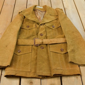 Puede incluir: Una chaqueta de ante color tostado con cuello ancho, mangas largas y un cinturón a juego con hebilla rectangular. La prenda incluye cuatro bolsillos delanteros con solapas abotonadas, cierre central de botones y un forro interior visible.