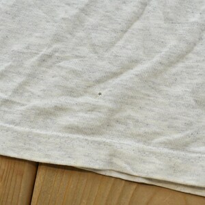 Op de afbeelding: Een lichtgrijs gem&ecirc;leerd t-shirt met een gespikkelde stoftextuur. Het kledingstuk heeft een zichtbare horizontale zoom aan de onderkant en een kleine, enkele donkere speldeprik-onvolkomenheid in het midden van het stofoppervlak.