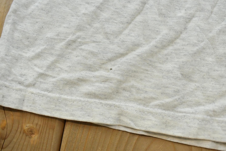 Op de afbeelding: Een lichtgrijs gem&ecirc;leerd t-shirt met een gespikkelde stoftextuur. Het kledingstuk heeft een zichtbare horizontale zoom aan de onderkant en een kleine, enkele donkere speldeprik-onvolkomenheid in het midden van het stofoppervlak.