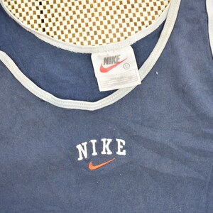 May include: A navy blue tank top with a white trim around the neck and armholes. The tank top has a white "NIKE" logo with an orange swoosh on the front.
