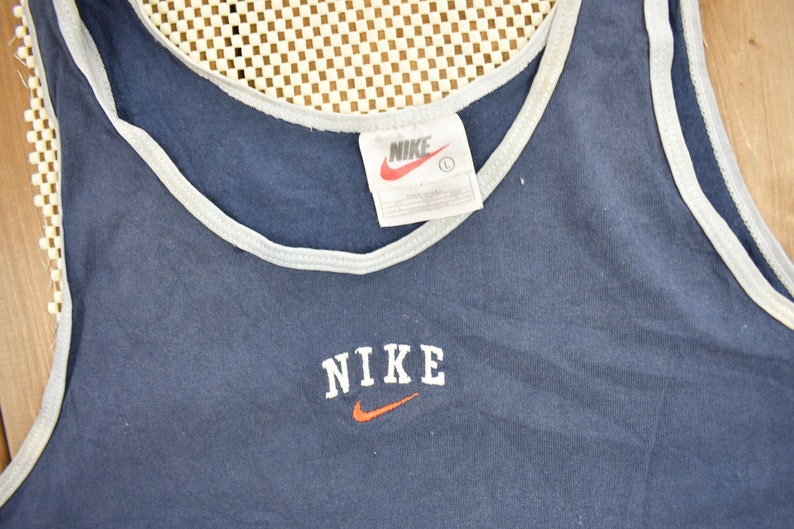 May include: A navy blue tank top with a white trim around the neck and armholes. The tank top has a white "NIKE" logo with an orange swoosh on the front.