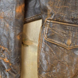 May include: Close-up of a distressed brown leather jacket. The jacket features a pocket and a zipper. The leather shows signs of wear with orange-brown discoloration. The jacket's texture is rough and aged, suggesting vintage style.