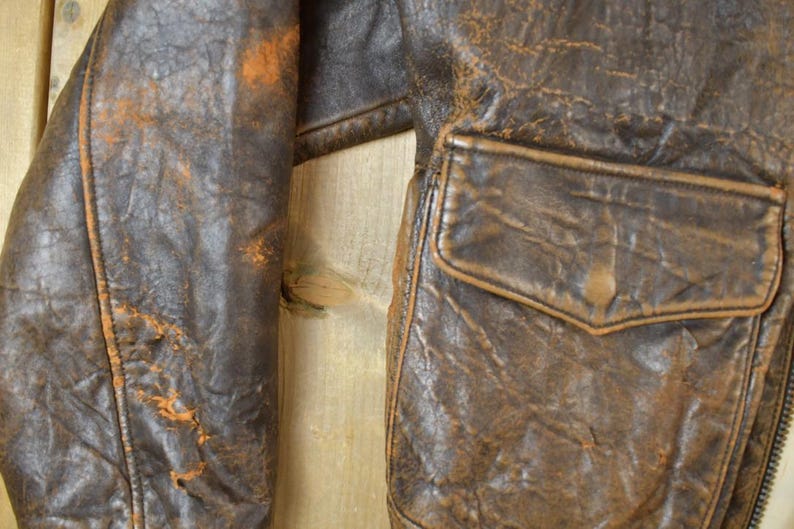 May include: Close-up of a distressed brown leather jacket. The jacket features a pocket and a zipper. The leather shows signs of wear with orange-brown discoloration. The jacket's texture is rough and aged, suggesting vintage style.