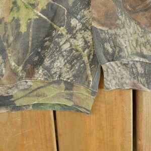 May include: A close-up of a camouflage-patterned sweatshirt. The fabric features a blend of green, brown, and gray hues, mimicking a forest environment. The ribbed cuffs and hem are visible, adding structure to the garment. The sweatshirt is made of a soft material.