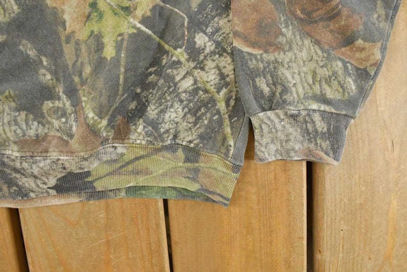 May include: A close-up of a camouflage-patterned sweatshirt. The fabric features a blend of green, brown, and gray hues, mimicking a forest environment. The ribbed cuffs and hem are visible, adding structure to the garment. The sweatshirt is made of a soft material.