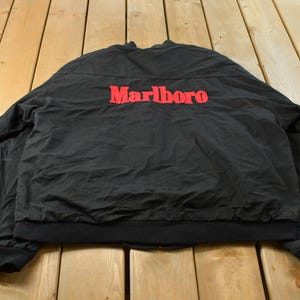 May include: A black bomber jacket featuring the word Marlboro embroidered in red lettering across the upper back. The jacket has long sleeves with black ribbed cuffs and a matching black ribbed hem at the waist, with a solid black fabric construction.