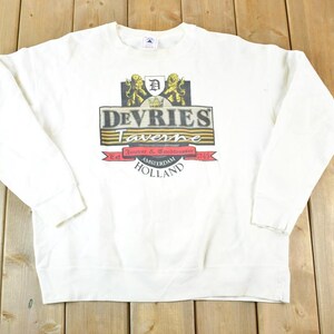 May include: Cream-coloured sweatshirt with a graphic that reads "Devries Taverne Amsterdam Holland." The graphic includes a crest with lions and a banner. The sweatshirt has long sleeves and a crew neck.