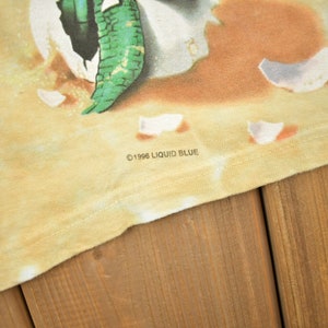 Puede incluir: Primer plano de una camiseta amarilla con un patr&oacute;n de tie-dye. La camiseta tiene un estampado gr&aacute;fico de una criatura verde que emerge de un huevo roto. El texto "&copy;1996 LIQUID BLUE" est&aacute; impreso en la parte inferior de la camiseta.