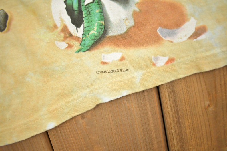 Puede incluir: Primer plano de una camiseta amarilla con un patr&oacute;n de tie-dye. La camiseta tiene un estampado gr&aacute;fico de una criatura verde que emerge de un huevo roto. El texto "&copy;1996 LIQUID BLUE" est&aacute; impreso en la parte inferior de la camiseta.