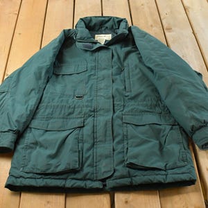 May include: A dark green insulated jacket featuring a high collar, a front zipper closure with a snap-button storm flap, two large lower patch pockets with flaps, two smaller chest pockets, and long sleeves with adjustable cuffs for a secure fit.