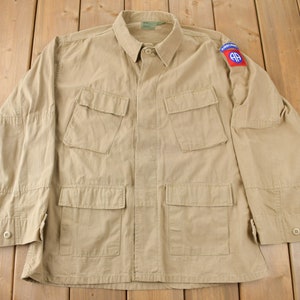 May include: A tan military-style jacket with four front pockets and a collared neckline. The jacket has a patch on the upper left sleeve with red, white, and blue accents. The jacket is buttoned and has buttoned cuffs.