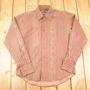 May include: A long-sleeved button-down shirt with a striped pattern in shades of red and white. The shirt features a collared neckline and buttoned cuffs. The fabric has a subtle textured appearance.