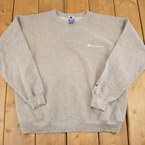 Peut inclure: Un sweat-shirt Champion gris chiné avec un col ras du cou et des manches longues. Le logo Champion est brodé en blanc sur la poitrine et un petit écusson Champion est sur la manche gauche. Le sweat-shirt est posé à plat sur une surface en bois.