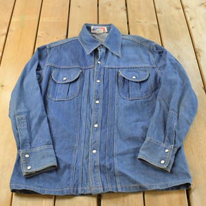 Puede incluir: Camisa vaquera vintage en un lavado azul medio, con cuello, cierre de botones y dos bolsillos en el pecho con solapas abotonadas. Las mangas largas tienen puños abotonados. La camisa está tendida sobre una superficie de madera.