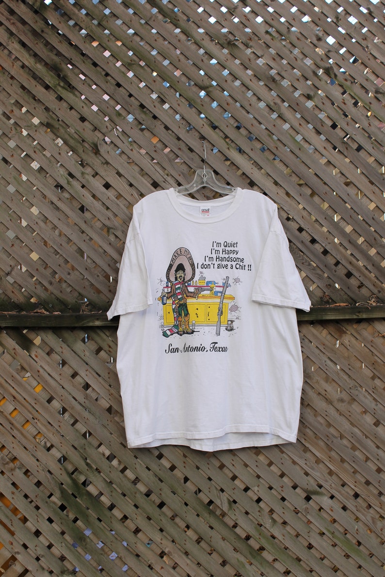 May include: White t-shirt with a cartoon illustration of a man wearing a sombrero and holding a bottle of beer. The text on the shirt reads "I'm Quiet I'm Happy I'm Handsome I don't give a Chit!!" and "San Antonio, Texas".
