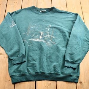 Op de afbeelding: Een tealgroen sweatshirt met lange mouwen met een witte grafische print van een winterscène. De afbeelding toont een besneeuwd landschap met bomen en een klein dier.