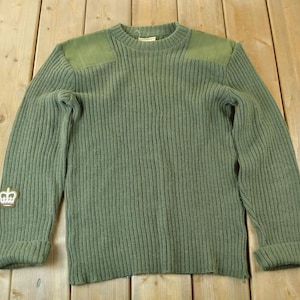 Może przedstawiać: Zielony sweter z dzianiny o prążkowanym splocie z naszywką z koroną na lewym rękawie. Sweter ma okrągły dekolt i jest wykonany z grubego, ciepłego materiału.