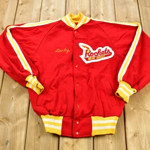 May include: Red and white varsity jacket with yellow stripes and a New Jersey Rockets patch. The jacket has a button closure and a name embroidered on the left chest: "Darby".