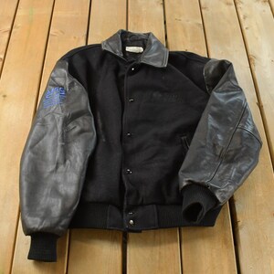 Könnte beinhalten: Schwarze Collegejacke mit grauen Lederärmeln und schwarzem geripptem Saum und Bündchen. Die Jacke hat eine Knopfleiste und einen grauen Lederkragen. Ein blauer Aufnäher ist auf dem linken Ärmel sichtbar.