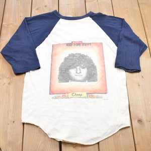 Puede incluir: Camiseta de béisbol blanca y azul marino con un gráfico vintage de la portada del álbum Kid for Rent. El gráfico presenta un retrato de una persona con cabello largo y el texto "Kid for Rent" y "Cheap".
