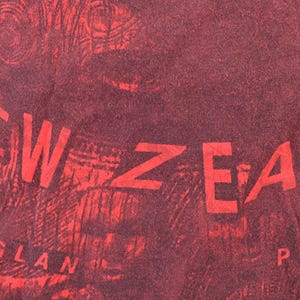 May include: Close-up of a red and maroon textile with the words "NEW ZEALAND" and "RAGLAN POINT" in a bold, sans-serif font. The background has a textured, almost wood-grain appearance.
