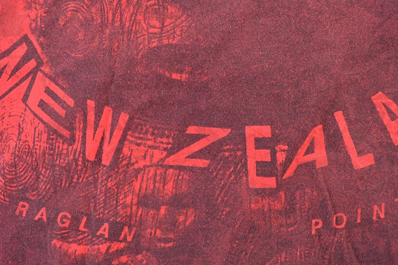 May include: Close-up of a red and maroon textile with the words "NEW ZEALAND" and "RAGLAN POINT" in a bold, sans-serif font. The background has a textured, almost wood-grain appearance.