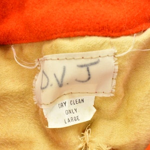 K&ouml;nnte beinhalten: Ein beigefarbenes Stofflabel mit dem Text "D.V.J." in schwarzer Schrift. Unter dem Text befindet sich ein kleineres wei&szlig;es Etikett mit dem Text "Dry Clean Only Large" in schwarzer Schrift.