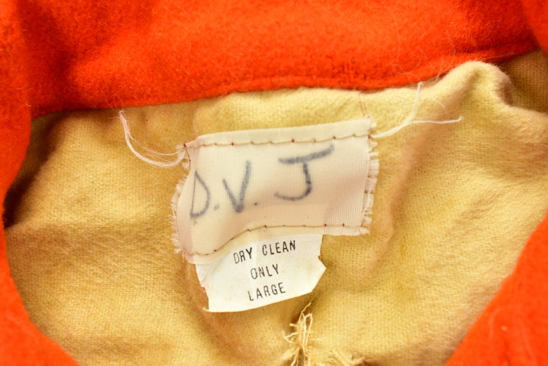 K&ouml;nnte beinhalten: Ein beigefarbenes Stofflabel mit dem Text "D.V.J." in schwarzer Schrift. Unter dem Text befindet sich ein kleineres wei&szlig;es Etikett mit dem Text "Dry Clean Only Large" in schwarzer Schrift.