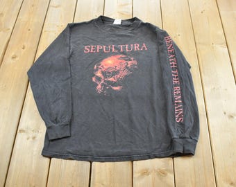 Puede incluir: Una camiseta negra de manga larga con el nombre de la banda SEPULTURA en texto rojo en el pecho sobre un gráfico rojo de una calavera. La manga izquierda tiene el texto BENEATH THE REMAINS impreso verticalmente en tinta roja a lo largo.