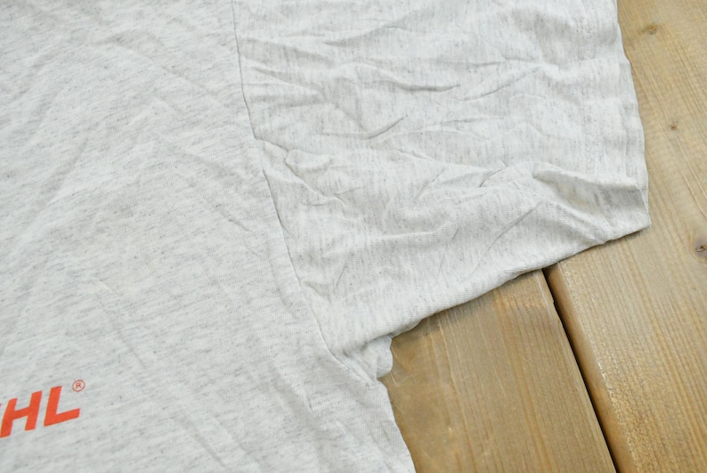 Op de afbeelding: Een lichtgrijs gem&ecirc;leerd T-shirt met de gedeeltelijke oranje tekst STIHL en een geregistreerd handelsmerksymbool zichtbaar aan de linkerkant. De stof heeft een getextureerd, gespikkeld uiterlijk met zichtbare vouwen en kreukels.