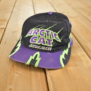 Op de afbeelding: Zwarte en paarse baseballcap met de tekst "ARCTIC CAT" in groen en paars. De cap heeft een gebogen klep en een knoop aan de bovenkant. De woorden "WORLD CLASS SNOWMOBILES" zijn ook zichtbaar.
