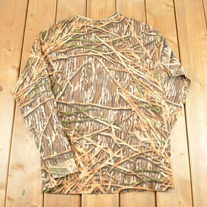 May include: Long-sleeved camouflage shirt in shades of brown, tan, and green. The shirt features a pattern designed to blend with natural environments. The garment is made of a lightweight material and is suitable for outdoor activities.