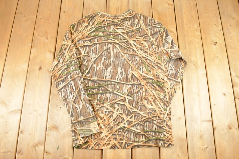 May include: Long-sleeved camouflage shirt in shades of brown, tan, and green. The shirt features a pattern designed to blend with natural environments. The garment is made of a lightweight material and is suitable for outdoor activities.