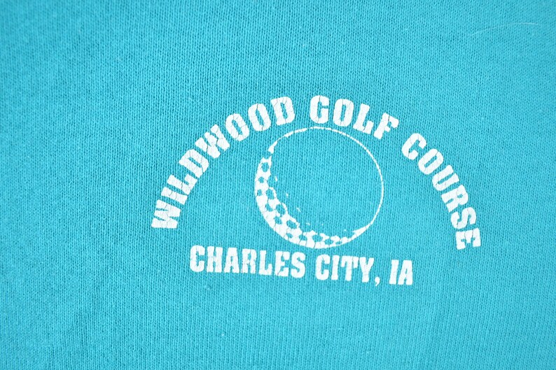 May include: Teal-colored fabric with a white graphic. The graphic features the words "Wildwood Golf Course" in an arch, with a golf ball design in the center. Below, the text reads "Charles City, IA."