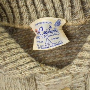 May include: Close-up of a beige wool sweater with a ribbed collar and a label that reads "Fine Virgin Wool" and "A Caldwell Hand Fashioned Canada". The sweater is made by Knit-Rite Mills Ltd.