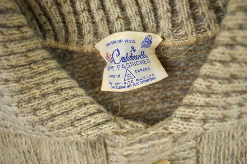 May include: Close-up of a beige wool sweater with a ribbed collar and a label that reads "Fine Virgin Wool" and "A Caldwell Hand Fashioned Canada". The sweater is made by Knit-Rite Mills Ltd.
