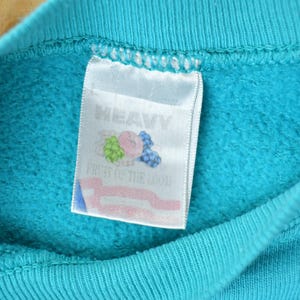 May include: Close-up of a turquoise blue sweatshirt collar with a white tag. The tag features the words "HEAVY" and "FRUIT OF THE LOOM" along with a graphic of a bunch of grapes, a pink peach, and blueberries.