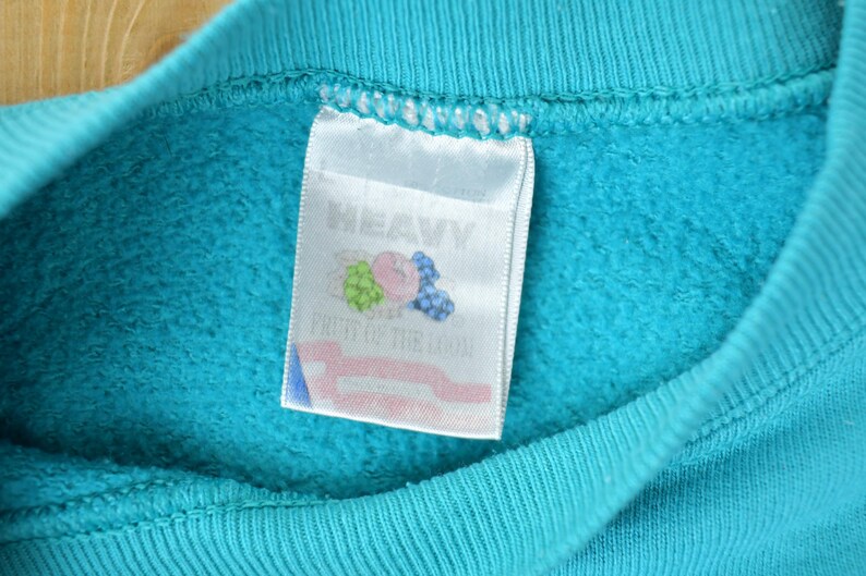 May include: Close-up of a turquoise blue sweatshirt collar with a white tag. The tag features the words "HEAVY" and "FRUIT OF THE LOOM" along with a graphic of a bunch of grapes, a pink peach, and blueberries.