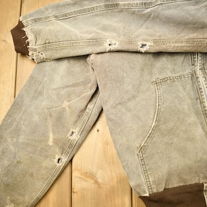 Może przedstawiać: Zużyta brązowa kurtka robocza z zamkiem błyskawicznym i naszywką z logo Carhartt. Kurtka ma kilka dziur i rozdarć, co świadczy o zużyciu.