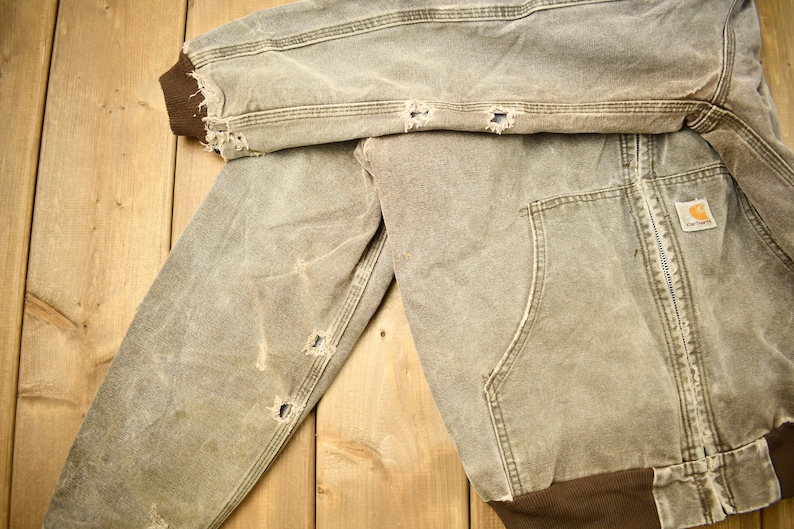 Może przedstawiać: Zużyta brązowa kurtka robocza z zamkiem błyskawicznym i naszywką z logo Carhartt. Kurtka ma kilka dziur i rozdarć, co świadczy o zużyciu.