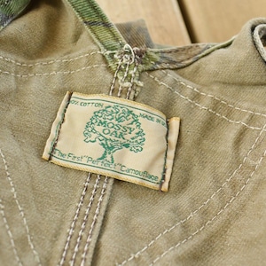 May include: Close-up of a tan and camouflage fabric with a "Mossy Oak" label. The label features a tree graphic and the text "The First 'Perfect' Camouflage". The fabric has visible stitching.