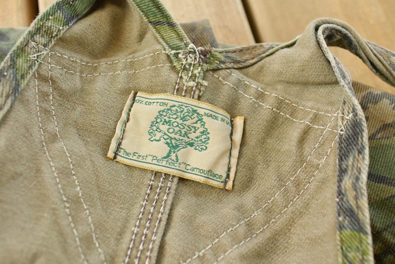May include: Close-up of a tan and camouflage fabric with a "Mossy Oak" label. The label features a tree graphic and the text "The First 'Perfect' Camouflage". The fabric has visible stitching.
